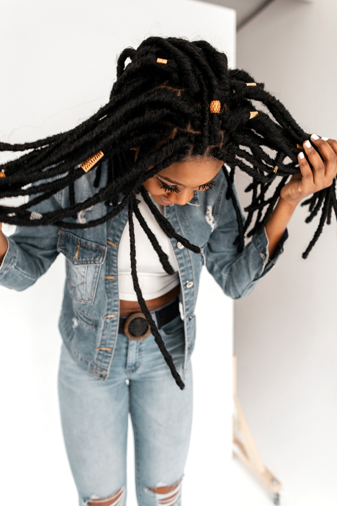 Pretty stylish modern black woman in fashion blue denim clothes looks down and straightens beautiful long dreadlocks in room.